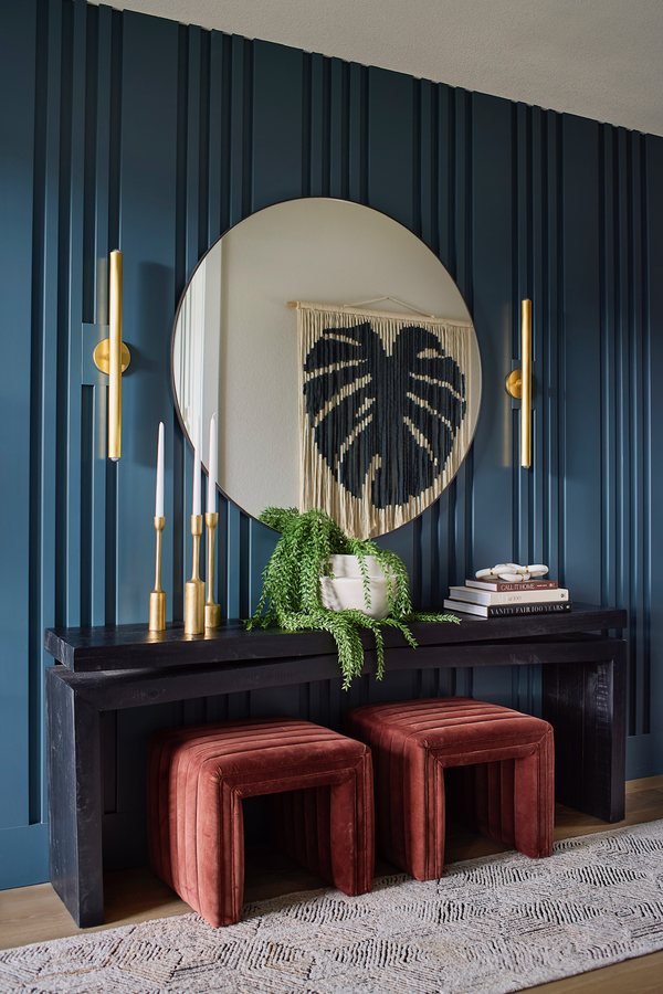 Round mirror, brass sconces and rust velvet ottomans against a navy slat-panel wall at the entry console in The Canyons new build, Castle Pines.